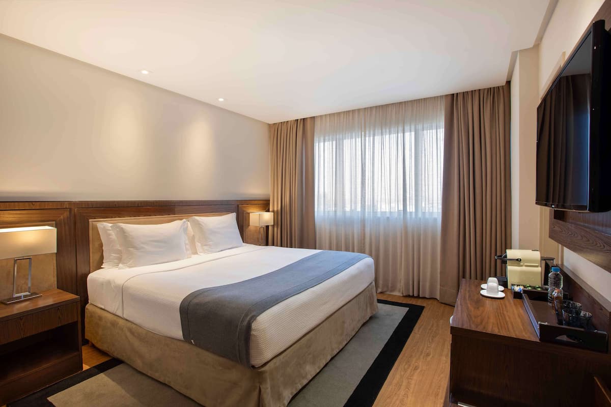 Una habitación de hotel con una cama grande, cabecero de madera, lámparas de noche, un televisor de pantalla plana y un escritorio con una silla.