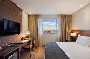 A hotel room with a bed, a desk, a chair, a television, and a window with a city view. - Windsor Brasilia Hotel (Brasília)