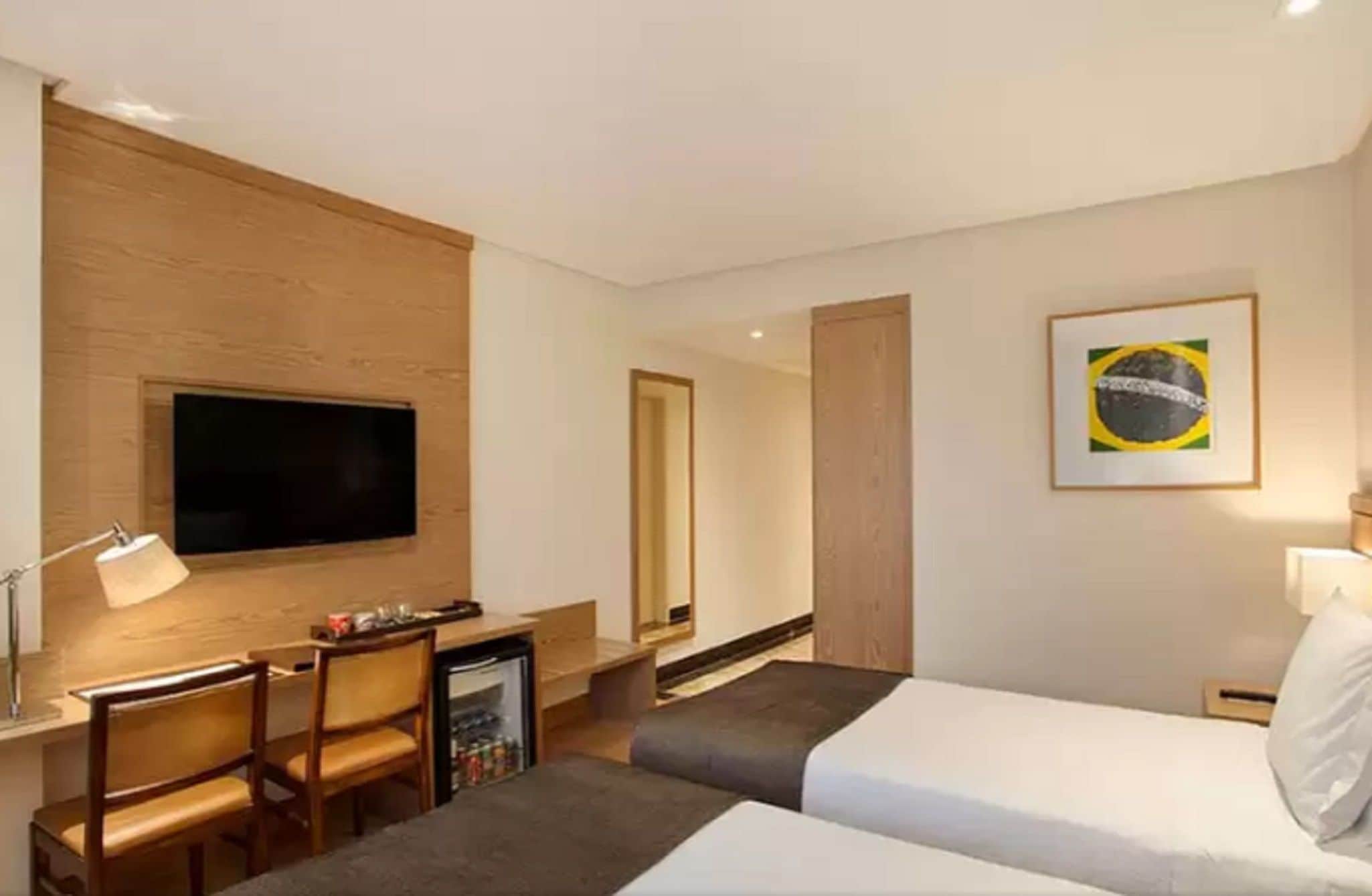 Una habitación de hotel con cama, escritorio con silla, televisor montado en la pared, refrigerador pequeño y un cuadro con la bandera brasileña.