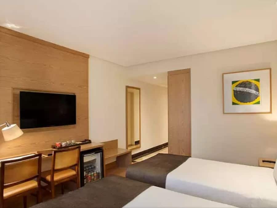 A hotel room with a bed, a desk with a chair, a television mounted on the wall, a small refrigerator, and a framed picture of the Brazilian flag.