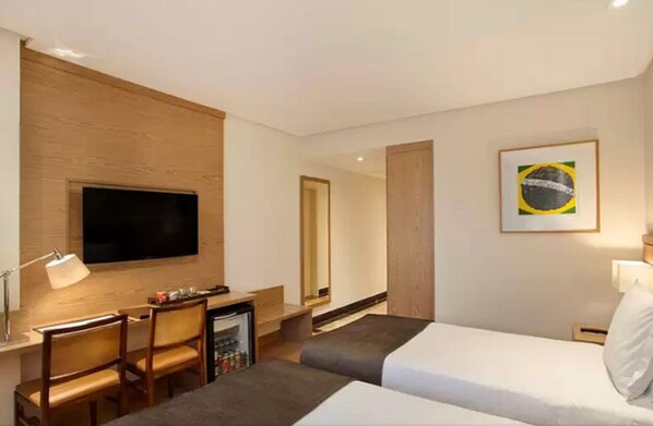 Une chambre d’hôtel comprenant un lit, un bureau avec une chaise, un téléviseur fixé au mur, un petit réfrigérateur et un tableau représentant le drapeau brésilien.
