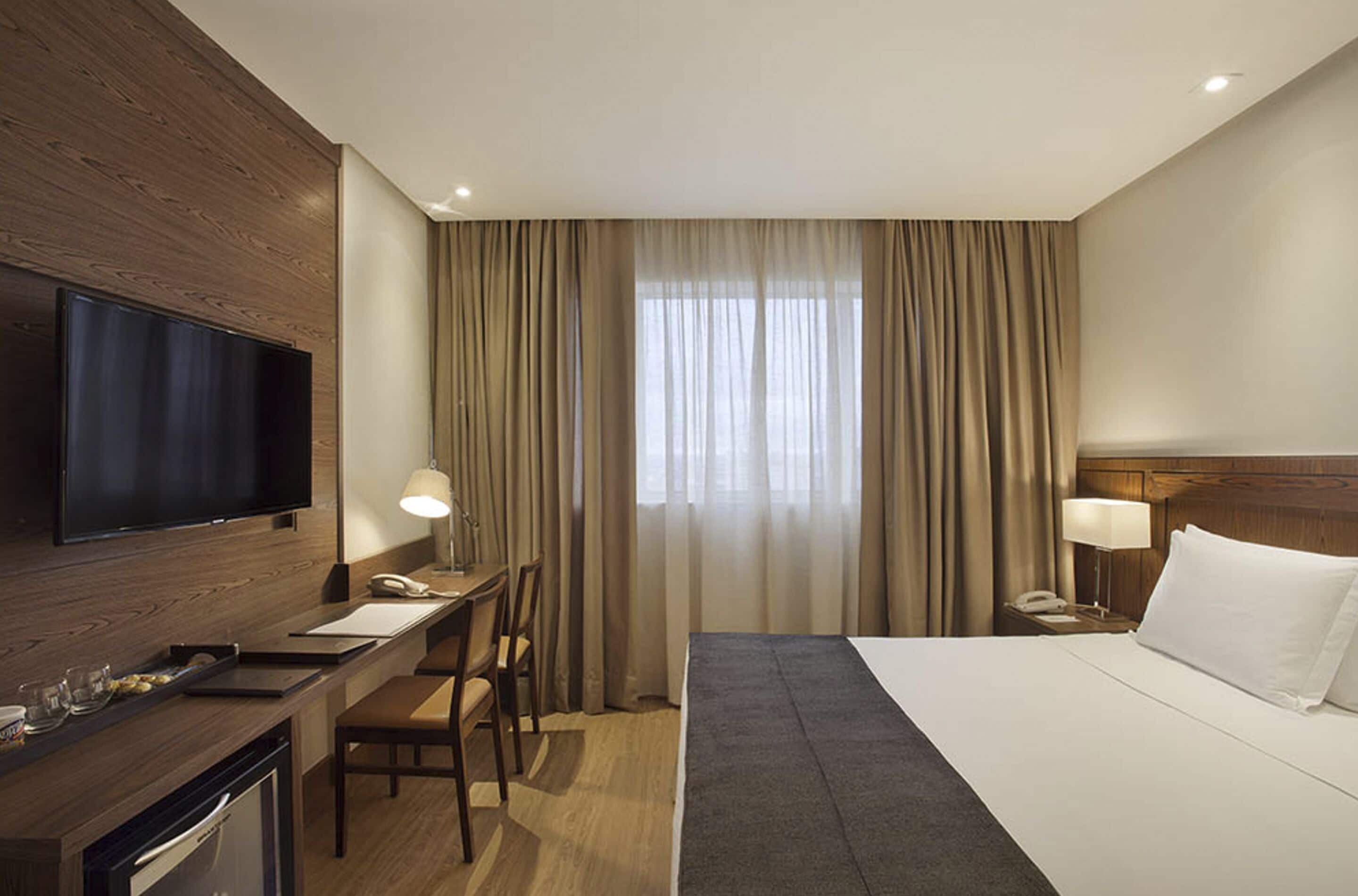 Habitación de hotel con cama, escritorio, televisión y ventana con cortinas.