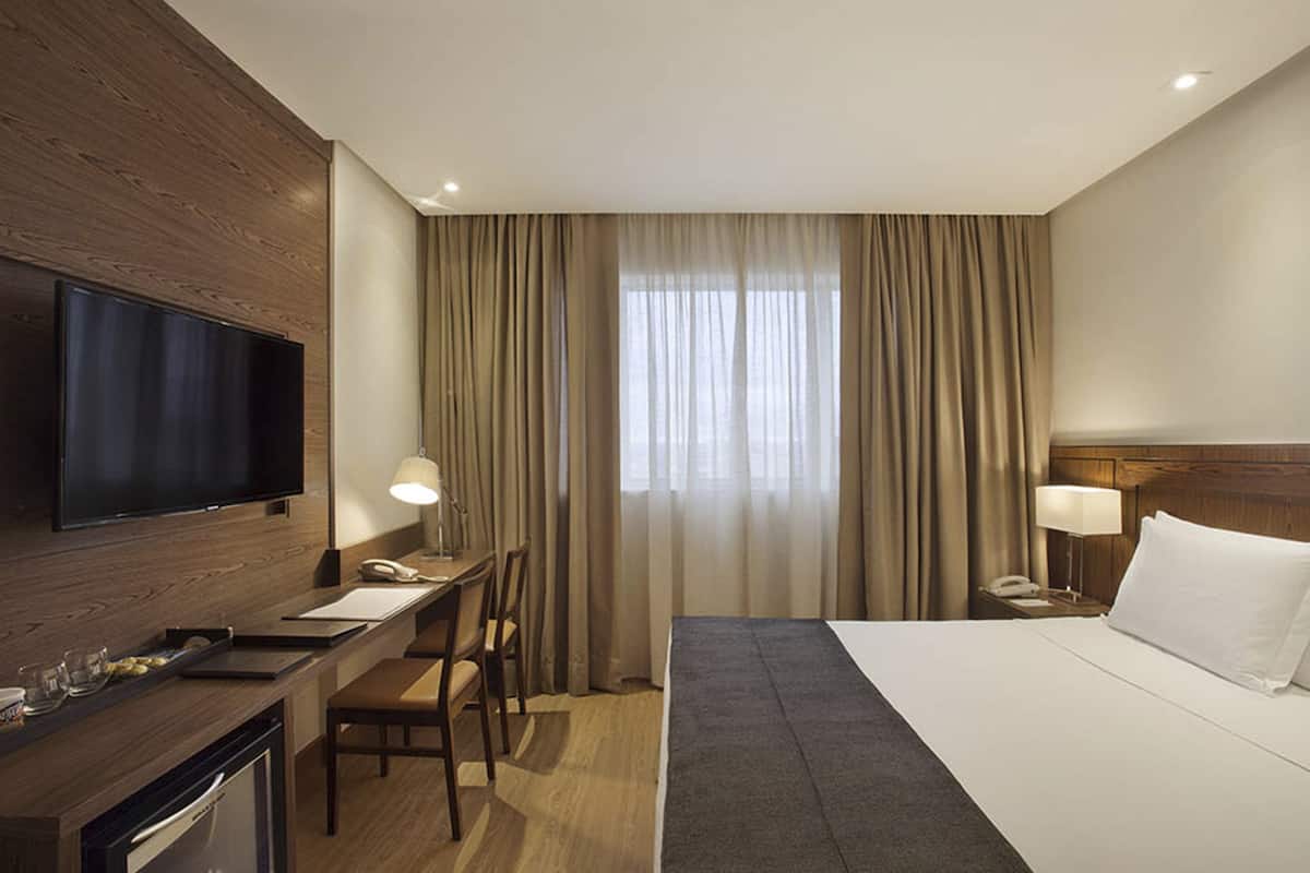 Habitación de hotel con cama, escritorio, televisión y ventana con cortinas.