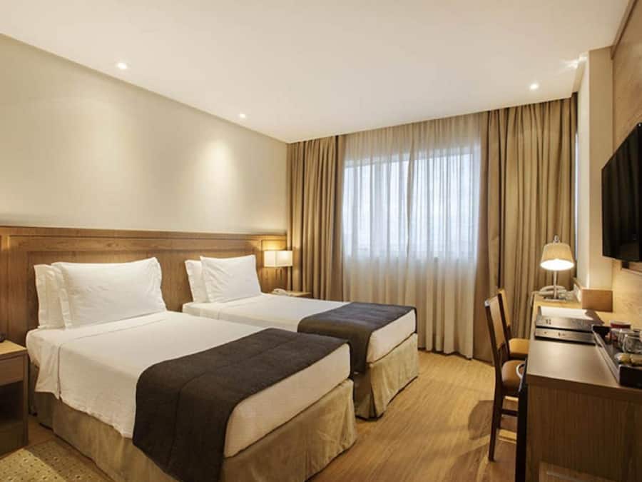Ein Hotelzimmer mit zwei Betten, einem Schreibtisch, einem Stuhl, einem Fernseher und einem Fenster mit Vorhängen.
