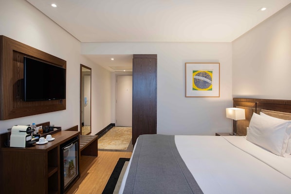 Quarto de hotel com cama, televisão, máquina de café, minibar e quadro na parede.