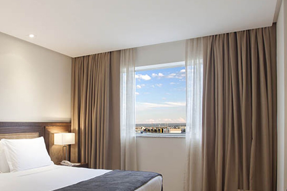 Ein Hotelzimmer mit einem Bett, einem Nachttisch mit Lampe und einem Fenster mit Vorhängen.