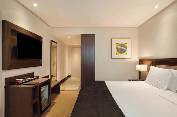 Ein Hotelzimmer mit einem Bett, einem Fernseher, einem Schreibtisch, einem Spiegel, einer Tür und einer Wand mit einem gerahmten Bild.
