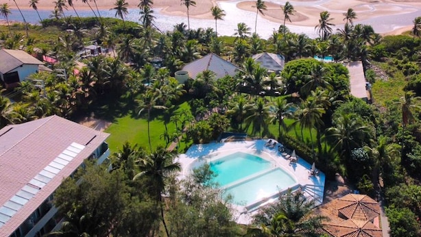 Vista aérea de un complejo turístico con una piscina central, rodeado de palmeras y una playa.
