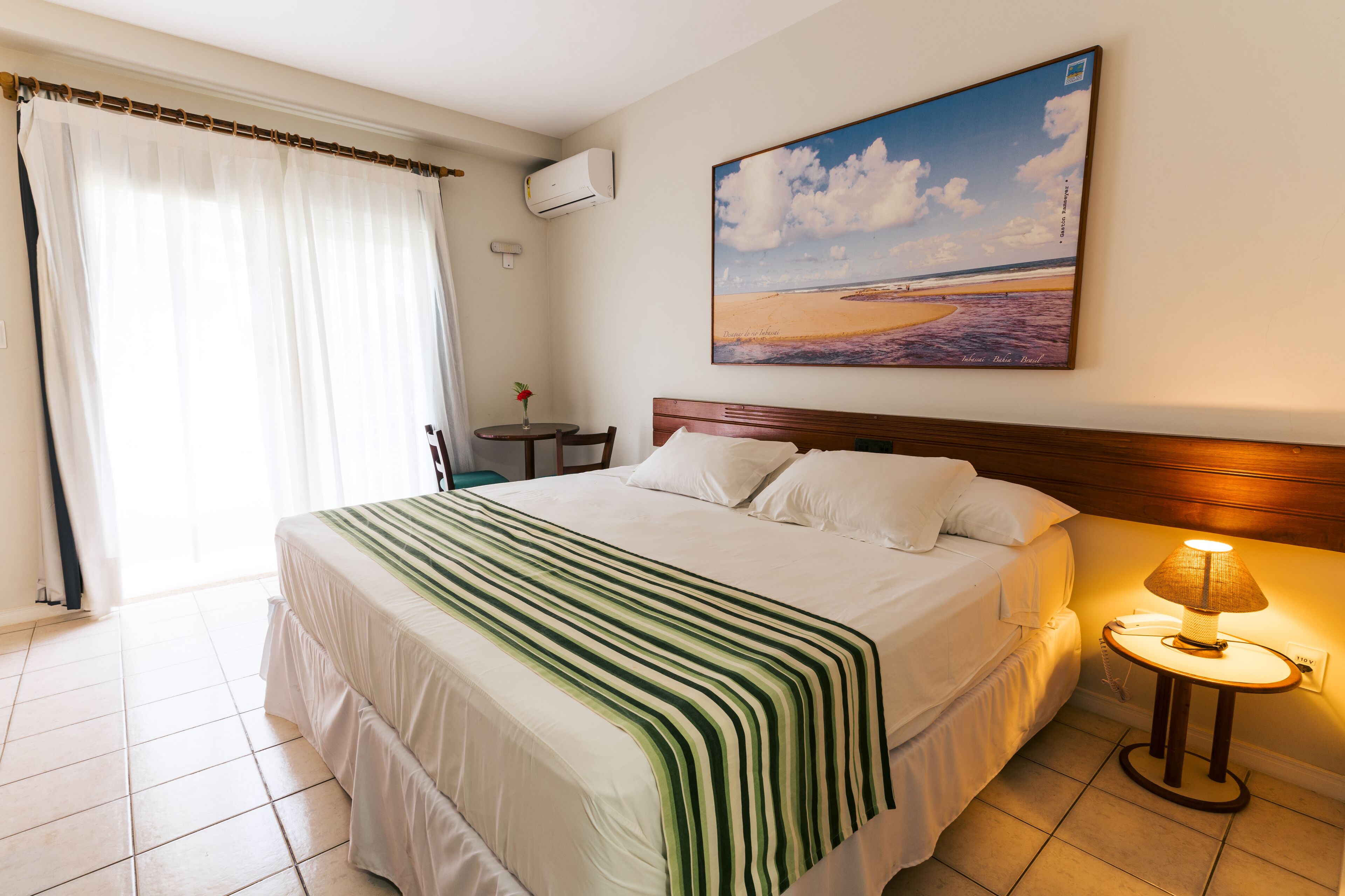 A hotel room with a bed, a bedside table with a lamp, a window with curtains, and a painting of a beach scene on the wall.