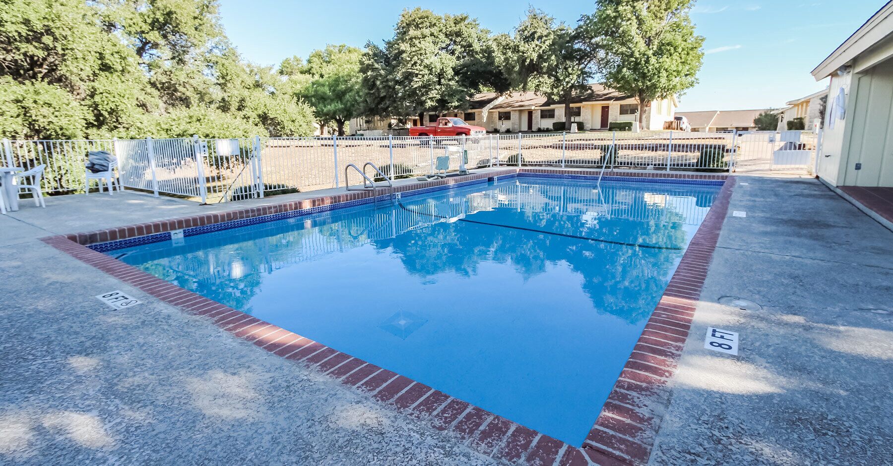 A rectangular swimming pool with a blue tiled border, surrounded by a white fence and trees in the background.