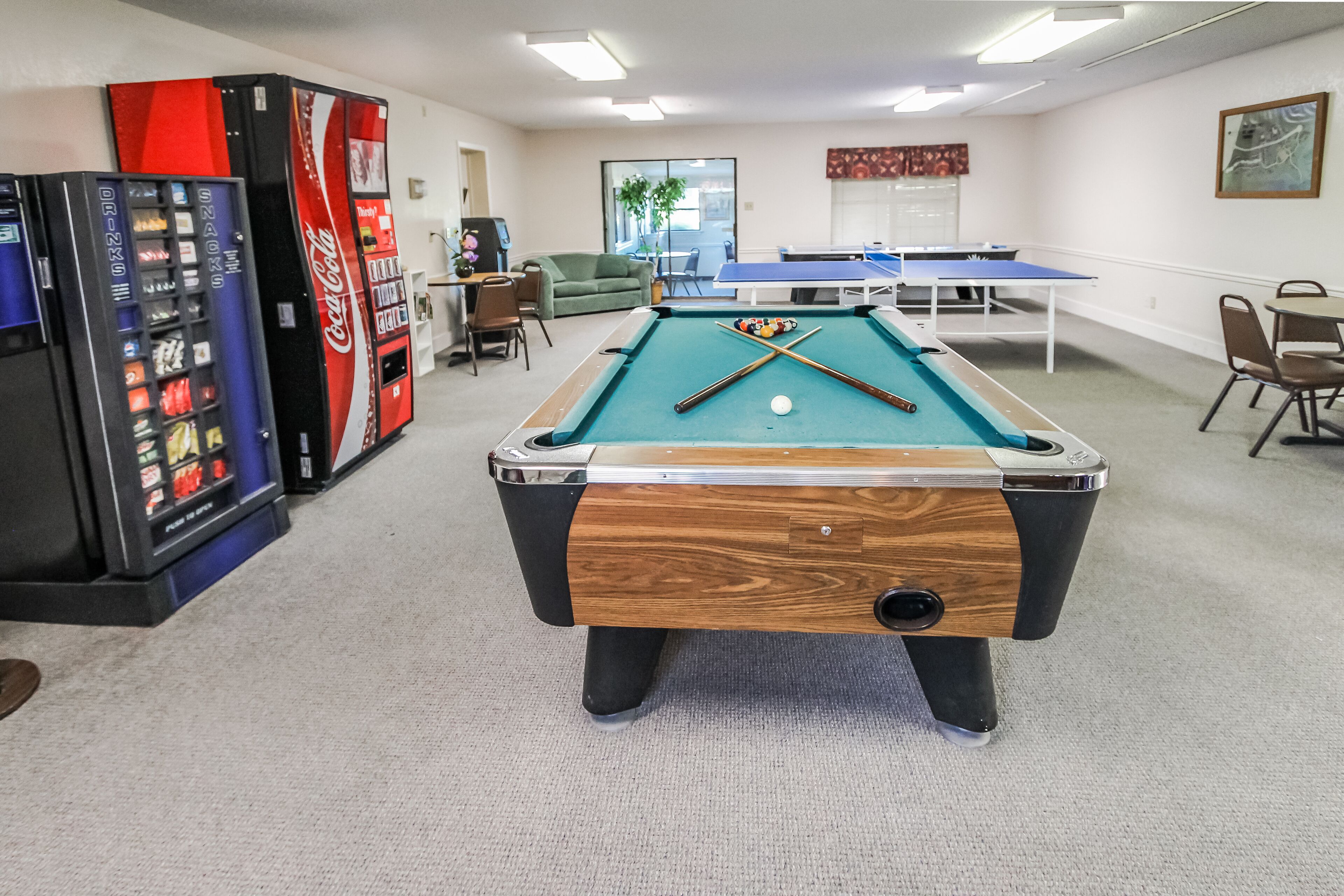 Una mesa de billar en una sala con una máquina expendedora de Coca-Cola, una mesa de ping pong y una pequeña zona de estar.