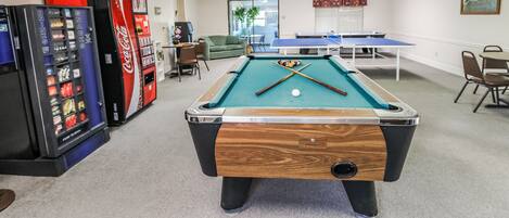 Una mesa de billar en una sala con una máquina expendedora de Coca-Cola, una mesa de ping pong y una pequeña zona de estar.