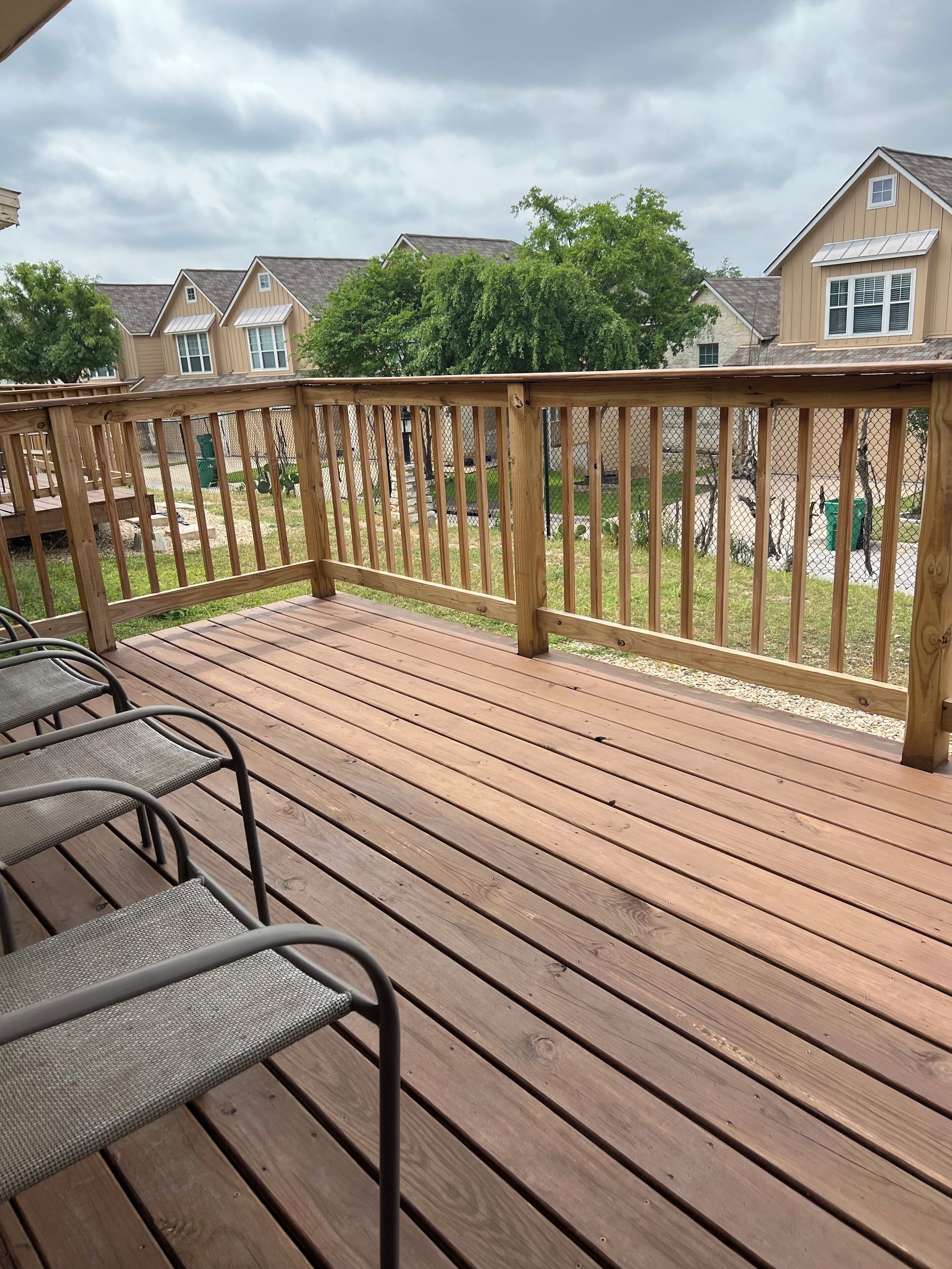 A wooden deck with metal chairs, surrounded by a chain-link fence and houses in the background.