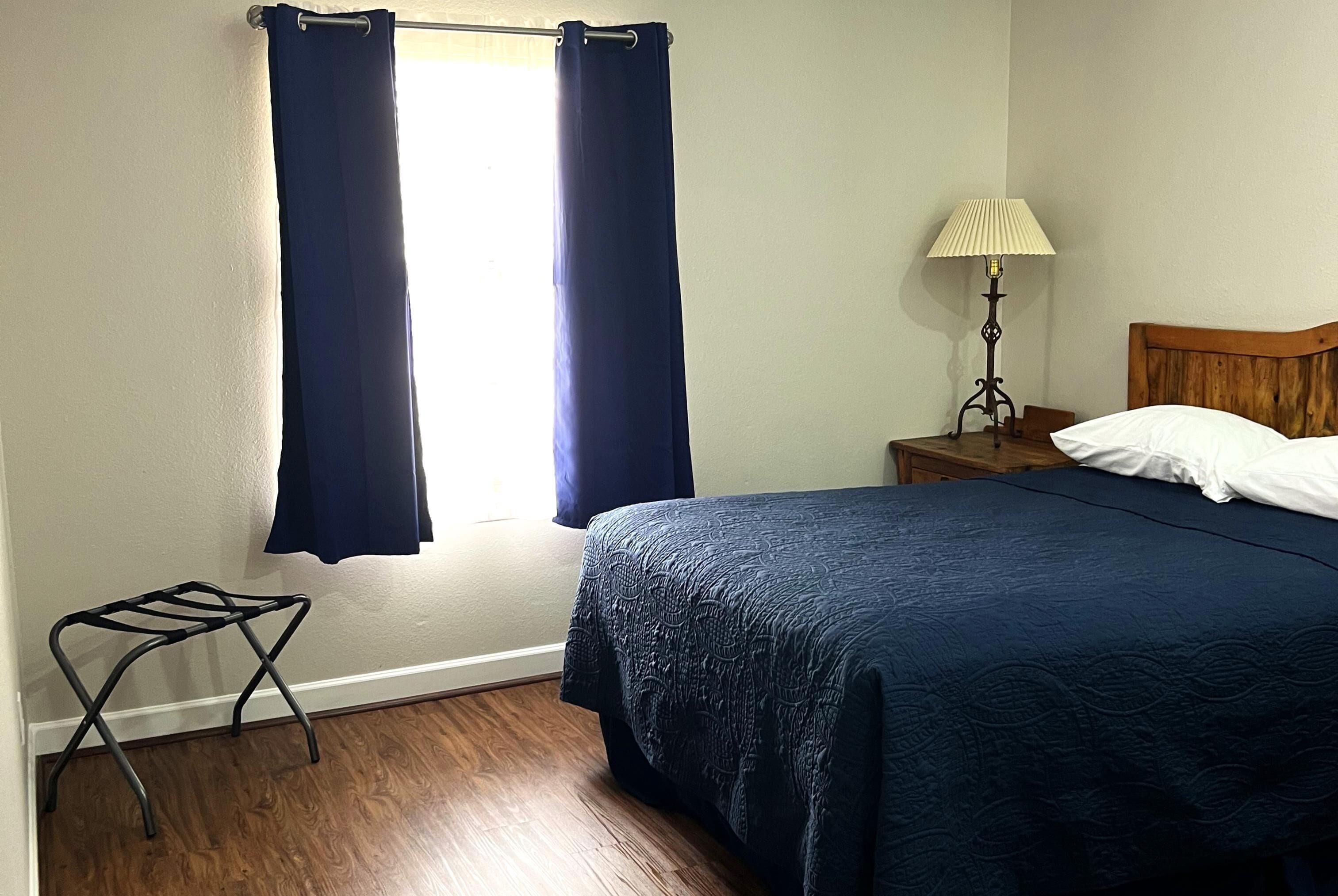 Un dormitorio con cama, una mesita de noche con lámpara, una ventana con cortinas y un taburete pequeño.