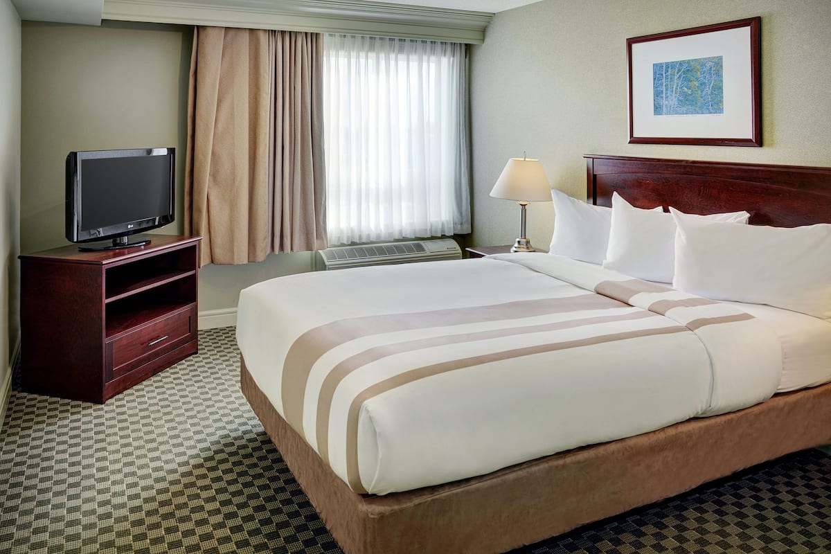 A hotel room with a large bed, a television on a wooden stand, an air conditioning unit, a lamp, and a framed picture on the wall.