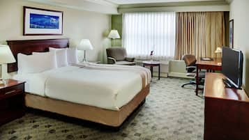 A hotel room with a large bed, two chairs, a desk with a TV, a lamp, and a painting on the wall.