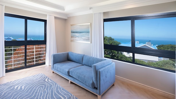 A modern living room with a blue sofa, large windows with a view of the sea, and a patterned rug. - Garden Route Hotel  (Mossel Bay)