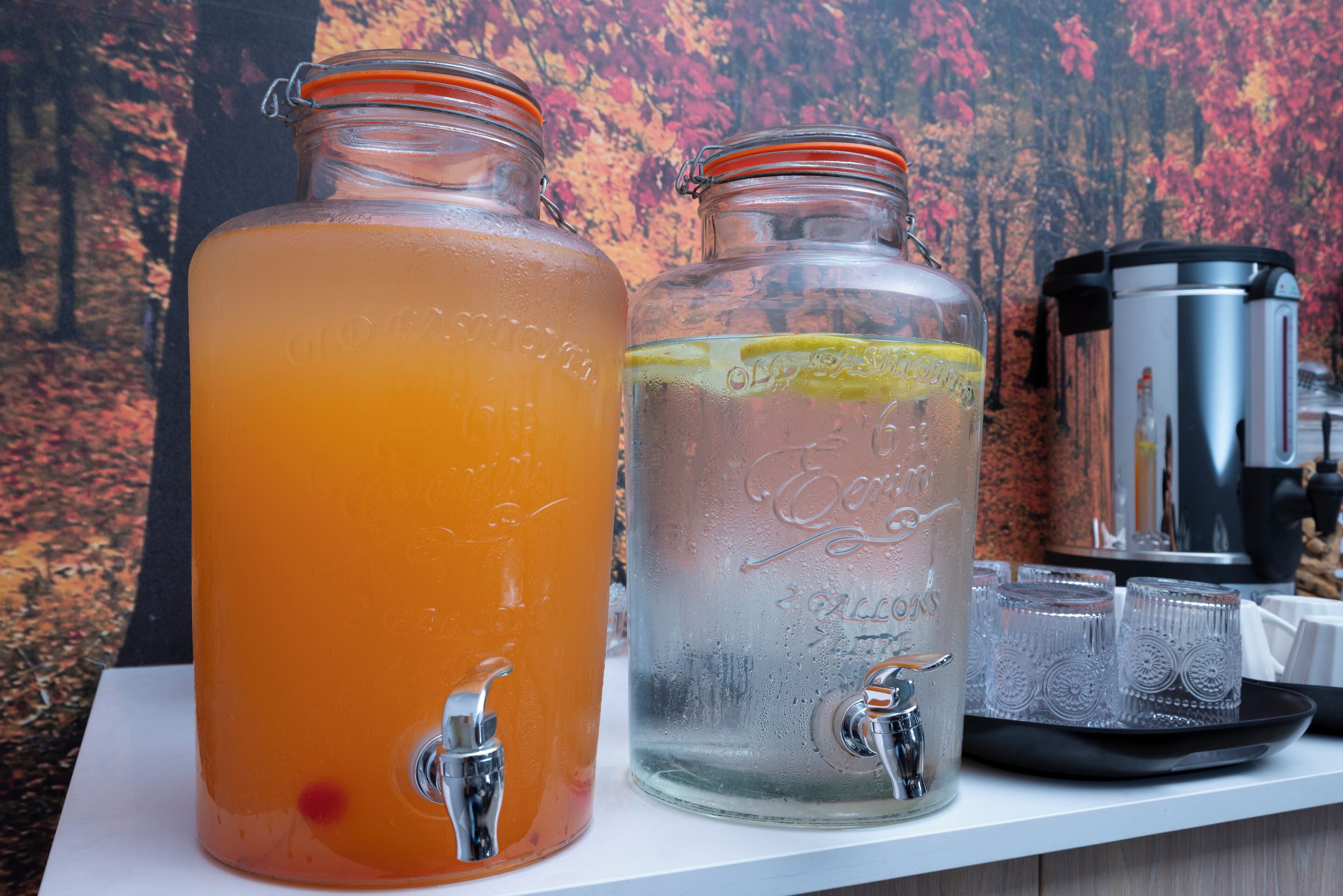 Two glass jars with taps, one filled with orange liquid and the other with clear liquid, and a coffee maker on a white surface.