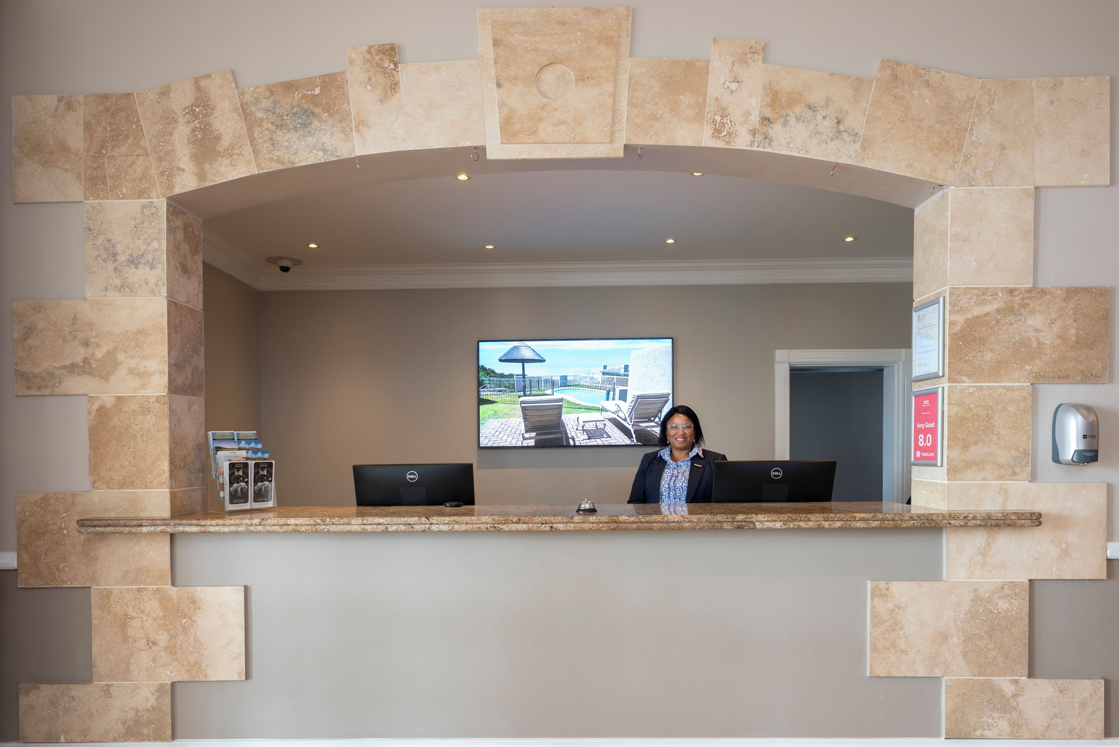 Un mostrador de recepción con un monitor que muestra una vista panorámica, dos computadoras portátiles y un cuadro en la pared.