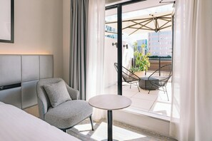 A modern hotel room with a bed, a grey armchair, a small round table, and a balcony with outdoor seating and a potted plant. - Hotel Centraza Hakata (Fukuoka)