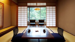 A traditional Japanese room with tatami flooring, a low table with two chairs, and a sliding door leading to a view of greenery.