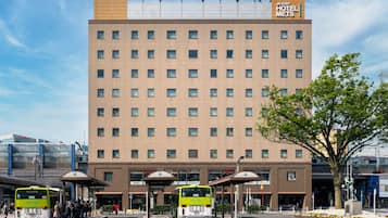 Edificio del Hotel Mets JR-East con varios pisos, una parada de autobús frente y una estatua en primer plano.