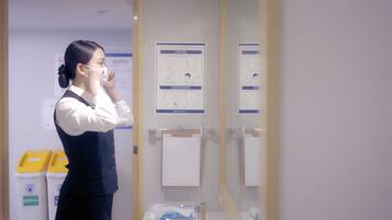 A person in a white shirt and dark vest standing in a room with a mirror, a door, and informational posters on the wall.