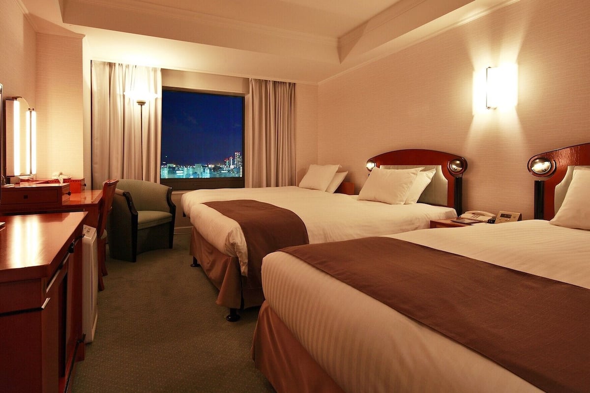 Ein Hotelzimmer mit zwei Betten, einem Schreibtisch, einem Stuhl, einem Fernseher und Blick auf die Stadt.