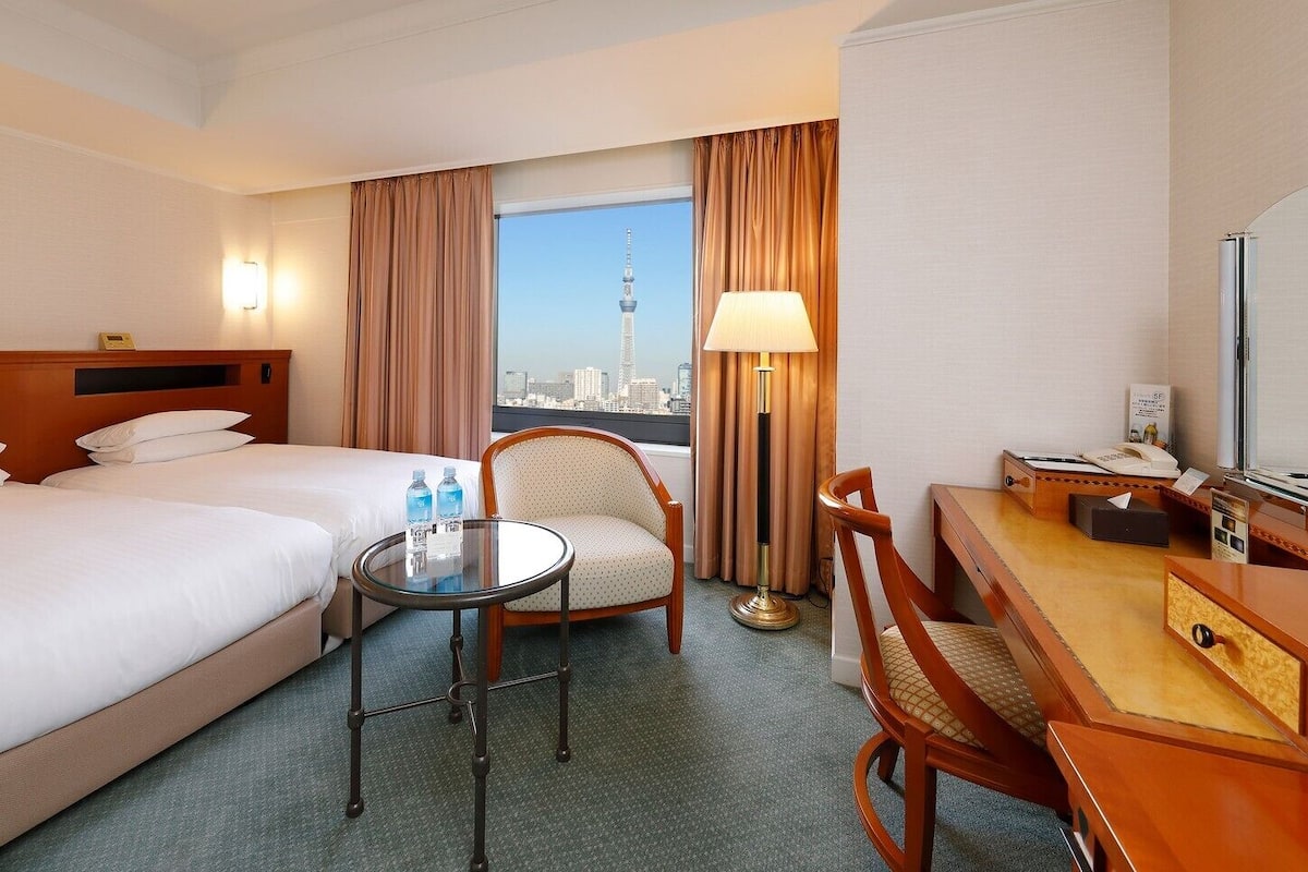 Ein Hotelzimmer mit zwei Betten, einem Schreibtisch, einem Stuhl, einem runden Tisch und Blick auf die Stadt.