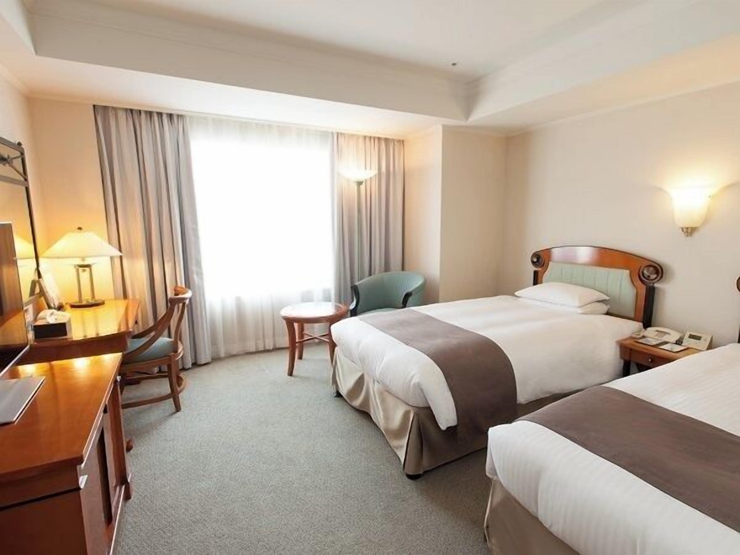 A hotel room with a bed, a desk, a chair, a small table, and a lamp.