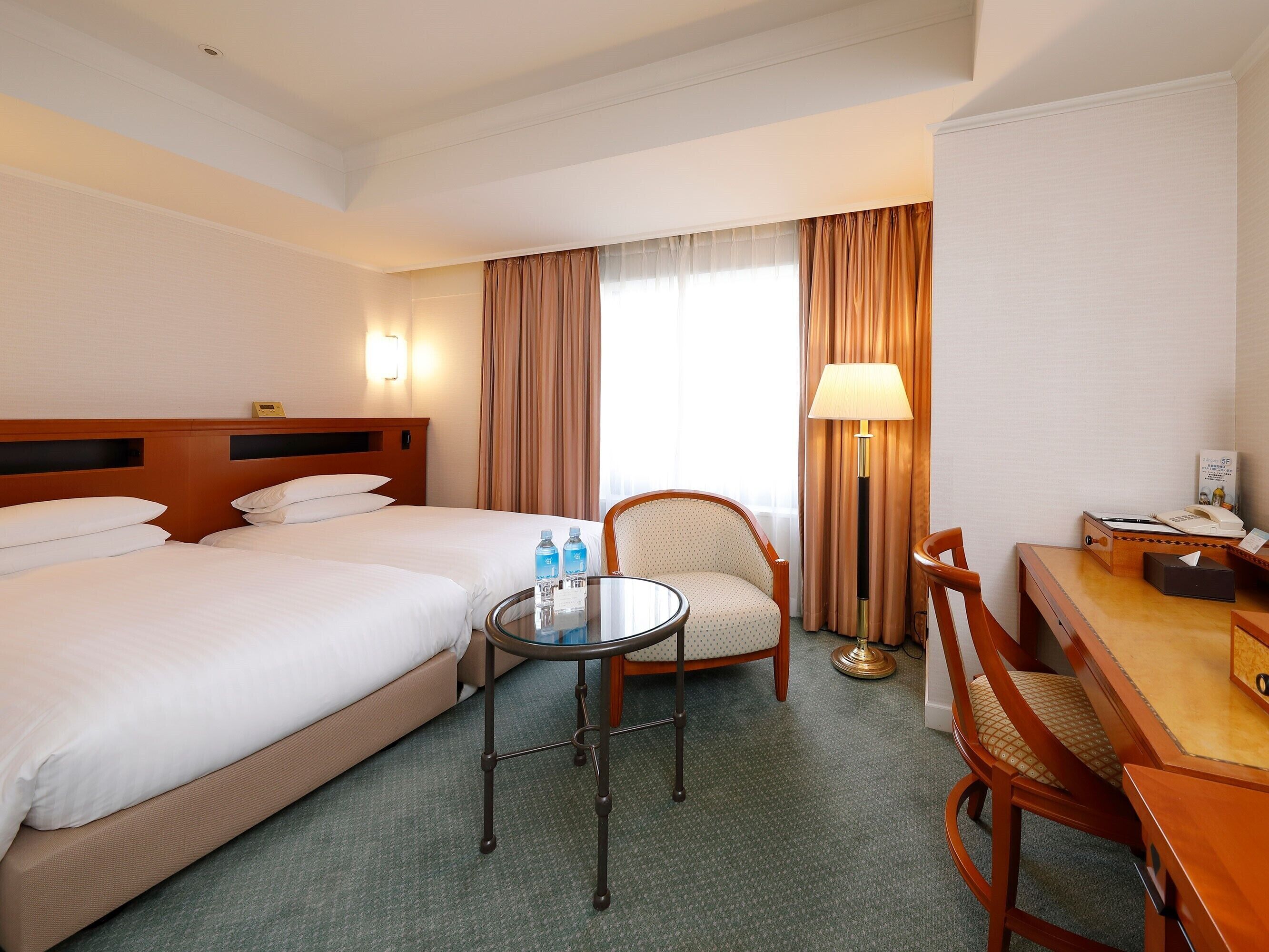 A hotel room with two beds, a desk with a telephone, a chair, a table, and a lamp.