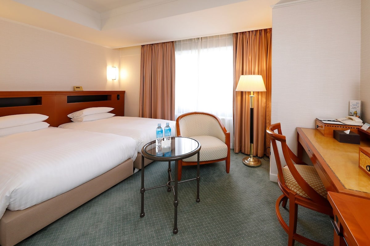 Ein Hotelzimmer mit zwei Betten, einem Schreibtisch mit Telefon, einem Sessel, einem Tisch und einer Lampe.