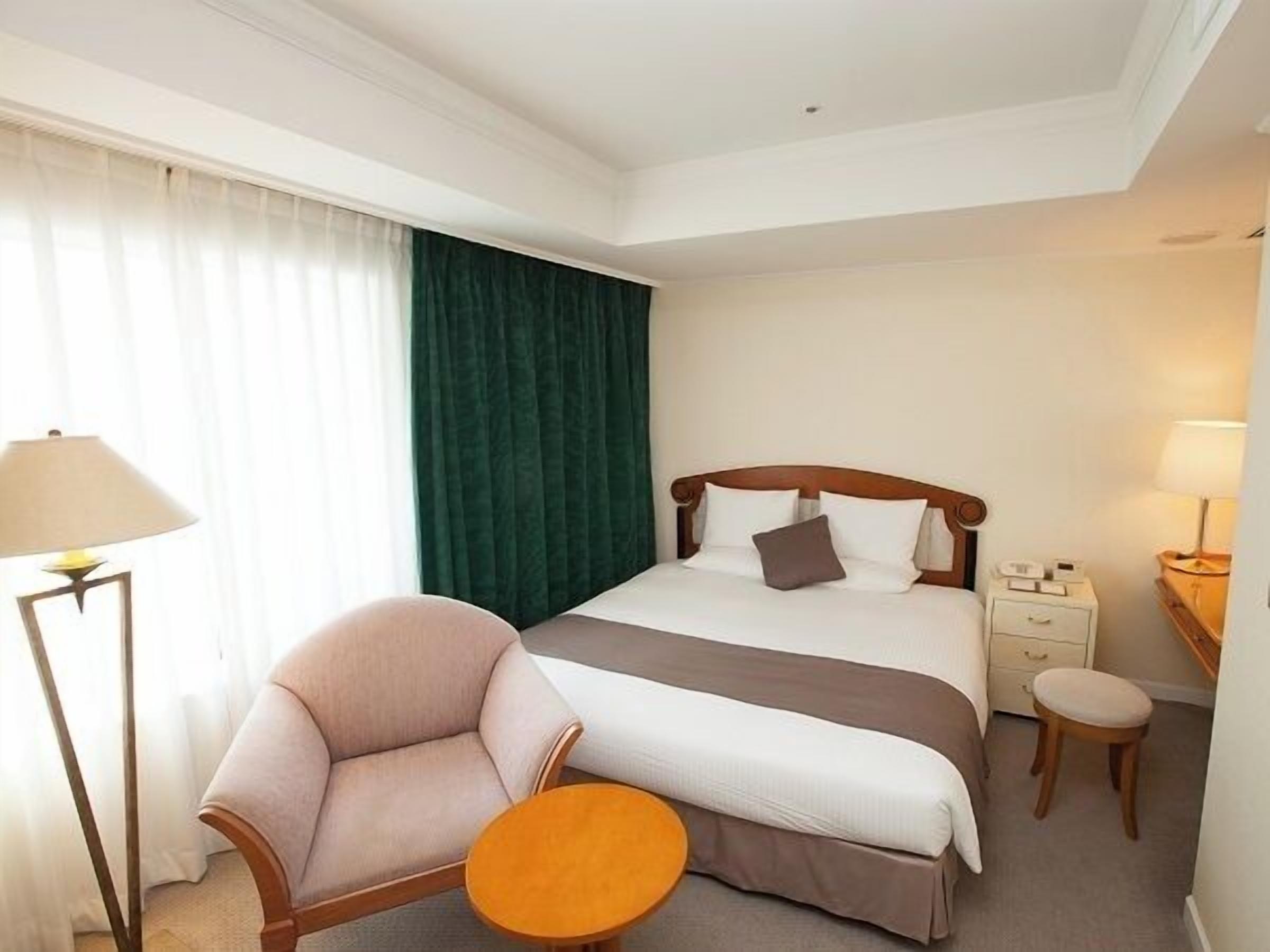A hotel room with a bed, a chair, a small table, a nightstand, and a lamp.
