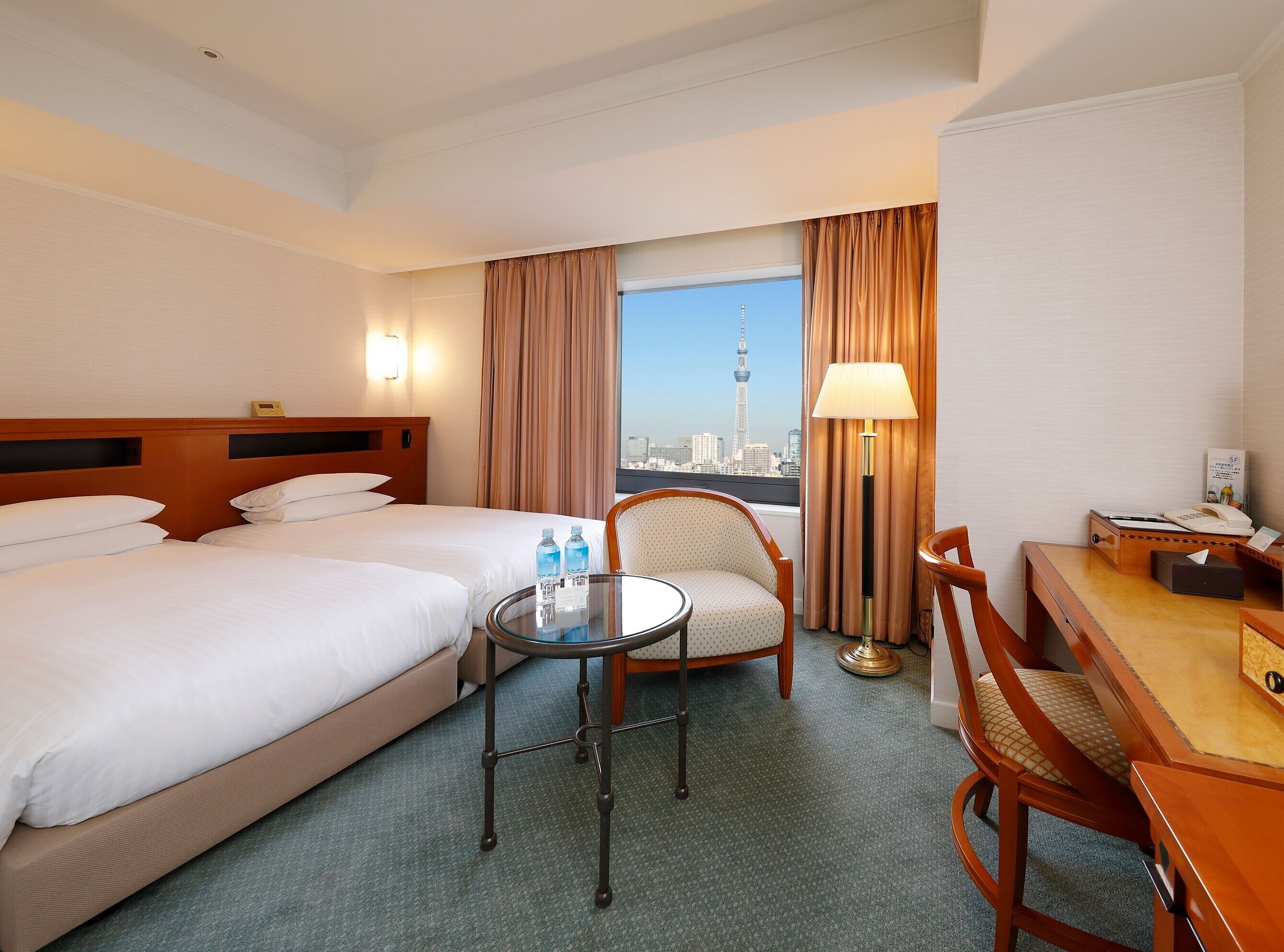A hotel room with two beds, a desk, a chair, a lamp, and a view of a cityscape.