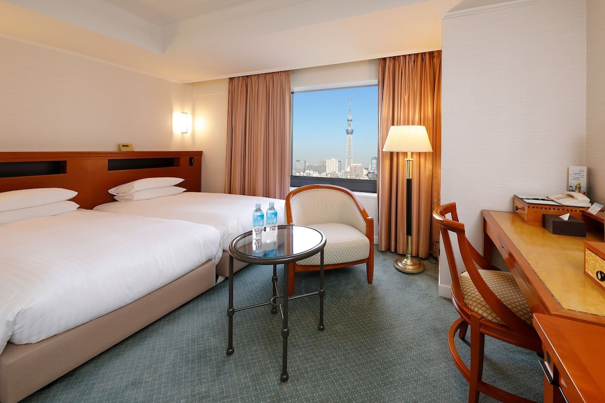 Ein Hotelzimmer mit zwei Betten, einem Schreibtisch, einem Stuhl, einer Lampe und Blick auf eine Stadtlandschaft.