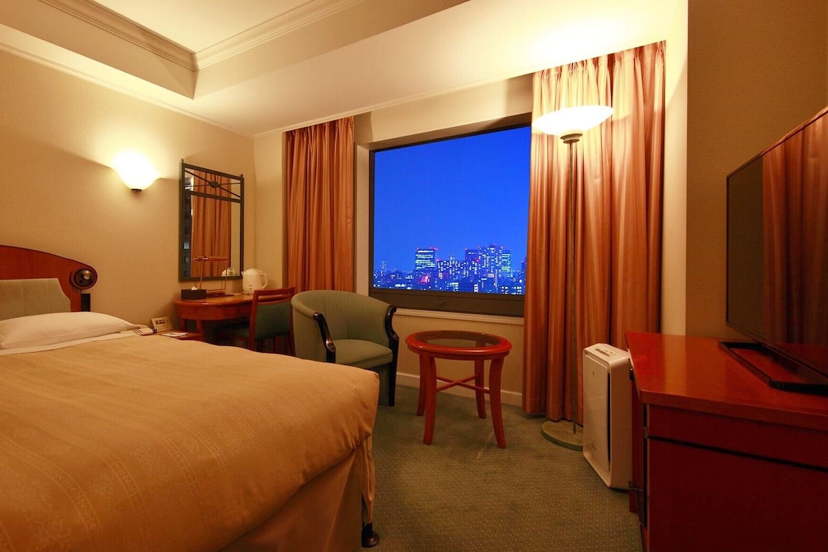 Ein Hotelzimmer mit einem Bett, einem Schreibtisch, einem Stuhl, einem Fernseher und einem Fenster mit Blick auf die Stadt.