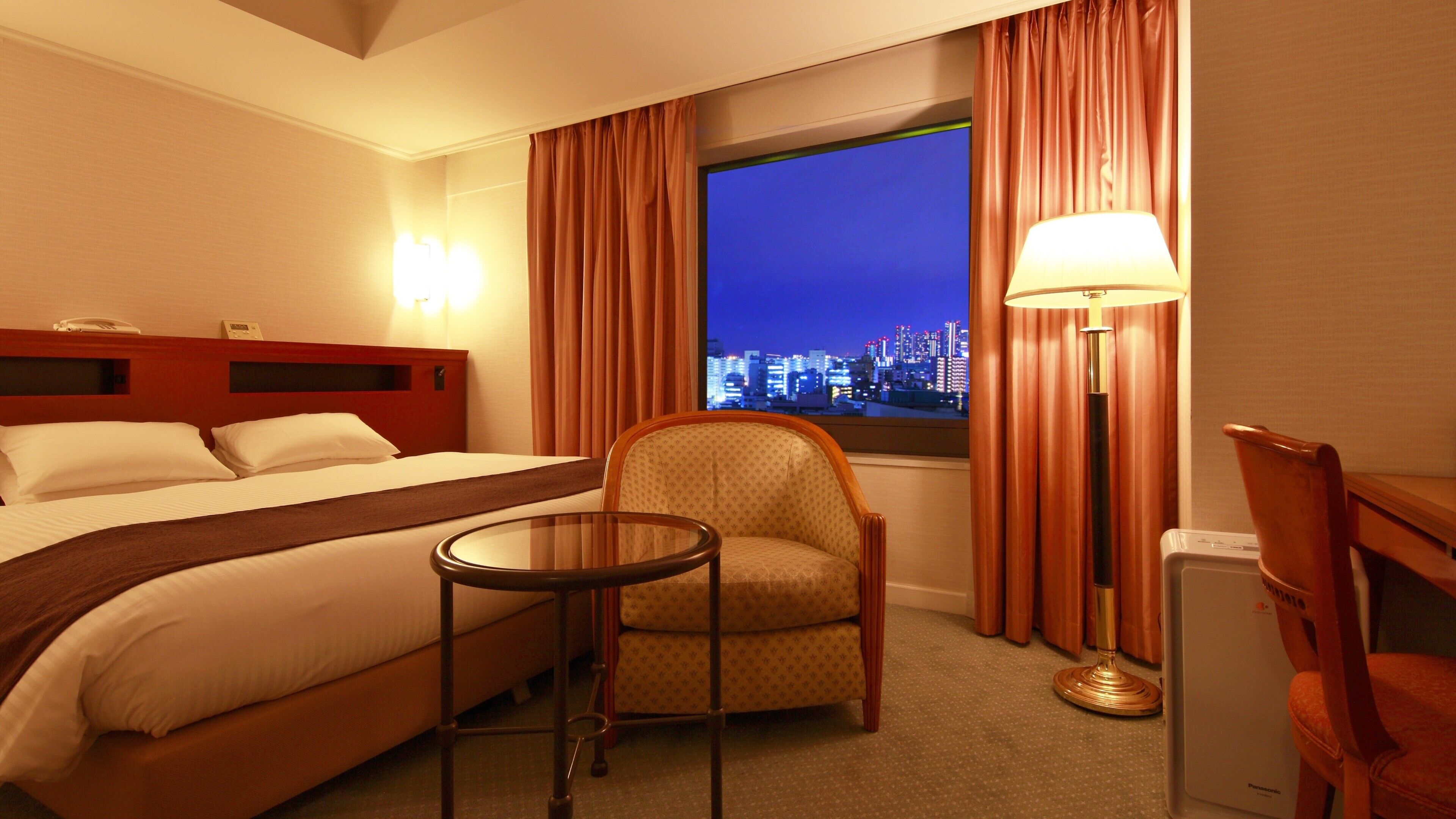 A hotel room with a bed, a chair, a table, a lamp, and a view of a cityscape at night.