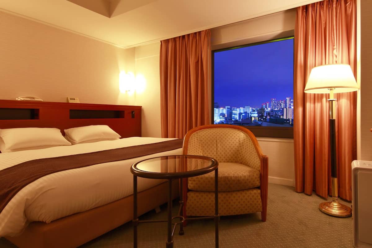 Ein Hotelzimmer mit Bett, Stuhl, Tisch, Lampe und Blick auf eine nächtliche Skyline.