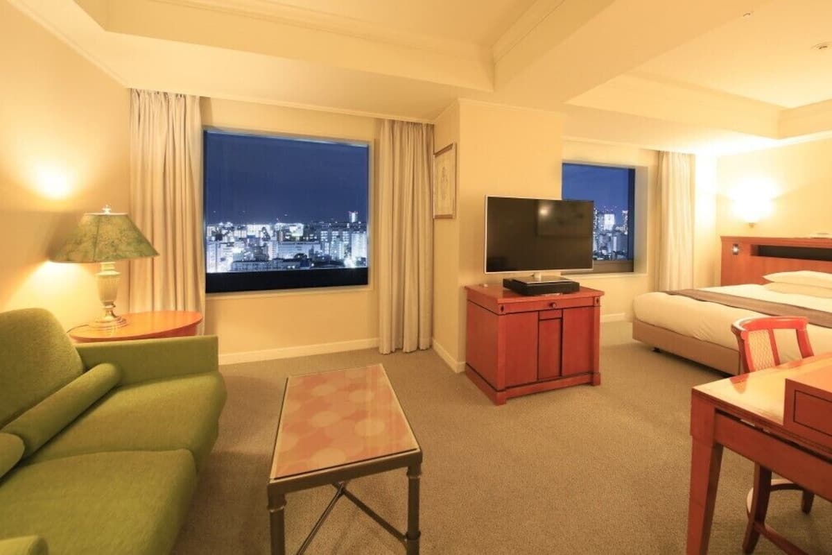 Ein Hotelzimmer mit Bett, Schreibtisch, Stuhl, Fernseher, Lampe und Blick auf nächtliche Stadtansicht.