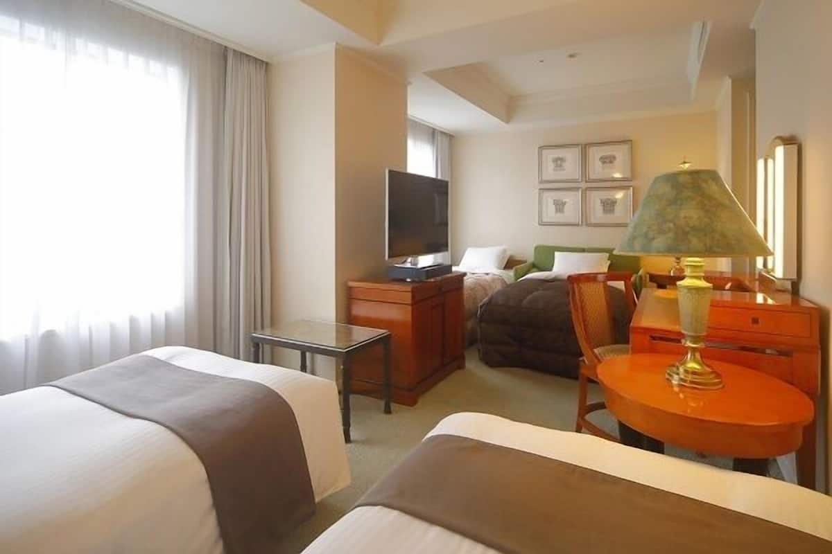 Ein Hotelzimmer mit zwei Betten, einem Fernseher, einem Schreibtisch, einem Stuhl, einer Lampe und gerahmten Bildern an der Wand.
