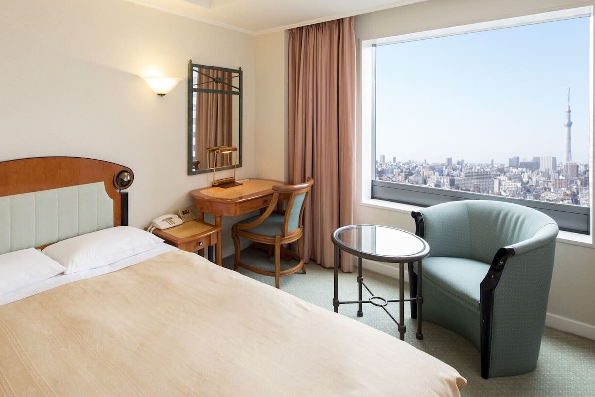 Ein Hotelzimmer mit einem Bett, einem Schreibtisch, einem Sessel, einem kleinen Tisch, einem Spiegel und einem Fenster mit Blick auf die Stadt.