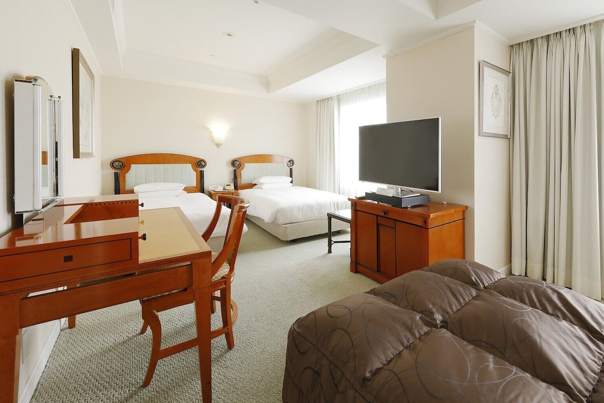 Ein Hotelzimmer mit zwei Betten, einem Schreibtisch, einem Stuhl, einem Fernseher und einem großen Fenster mit Vorhängen.