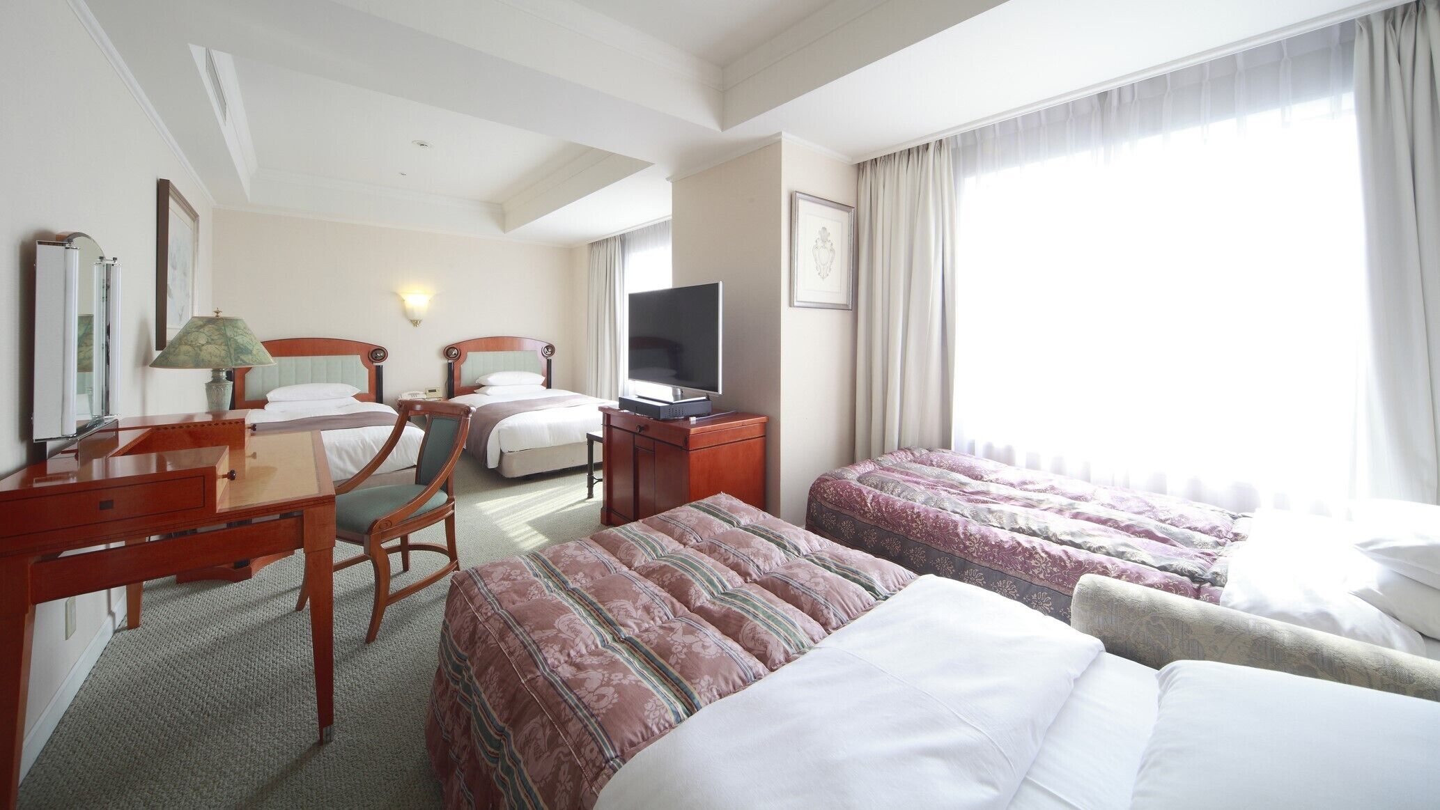 A hotel room with two beds, a desk with a lamp, a television, and a large window with curtains.