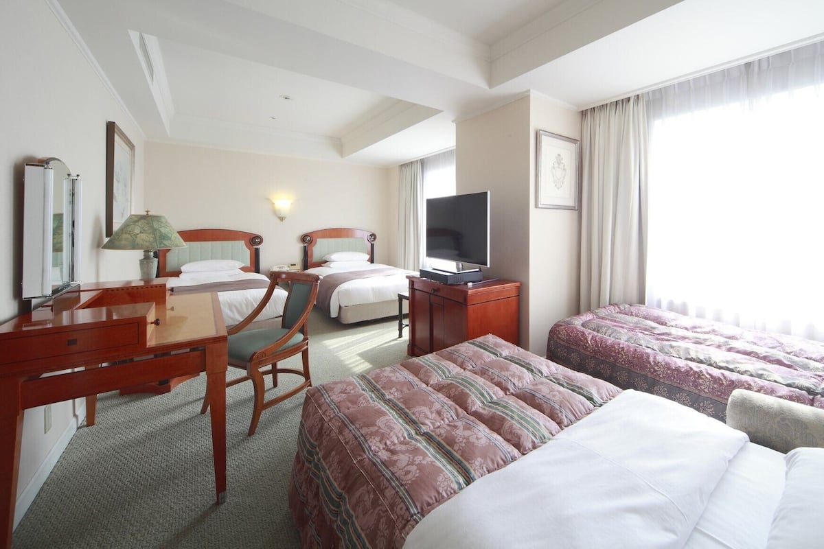 Ein Hotelzimmer mit zwei Betten, einem Schreibtisch mit Lampe, einem Fernseher und einem großen Fenster mit Vorhängen.