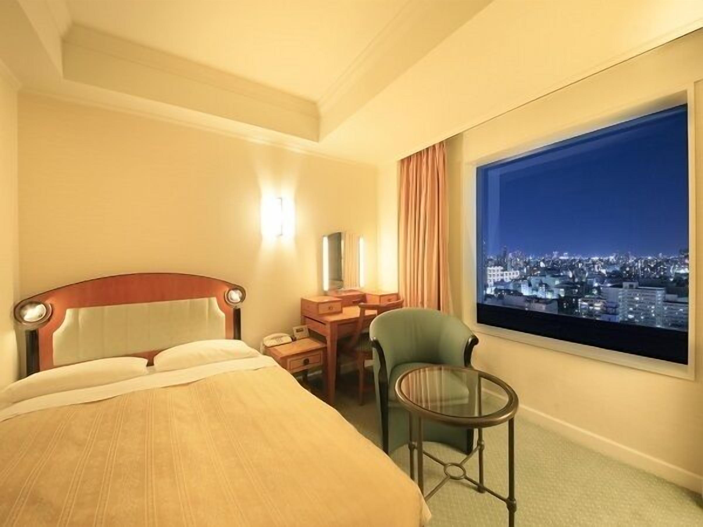 A hotel room with a bed, a desk, a chair, a small table, and a large window with a city view.