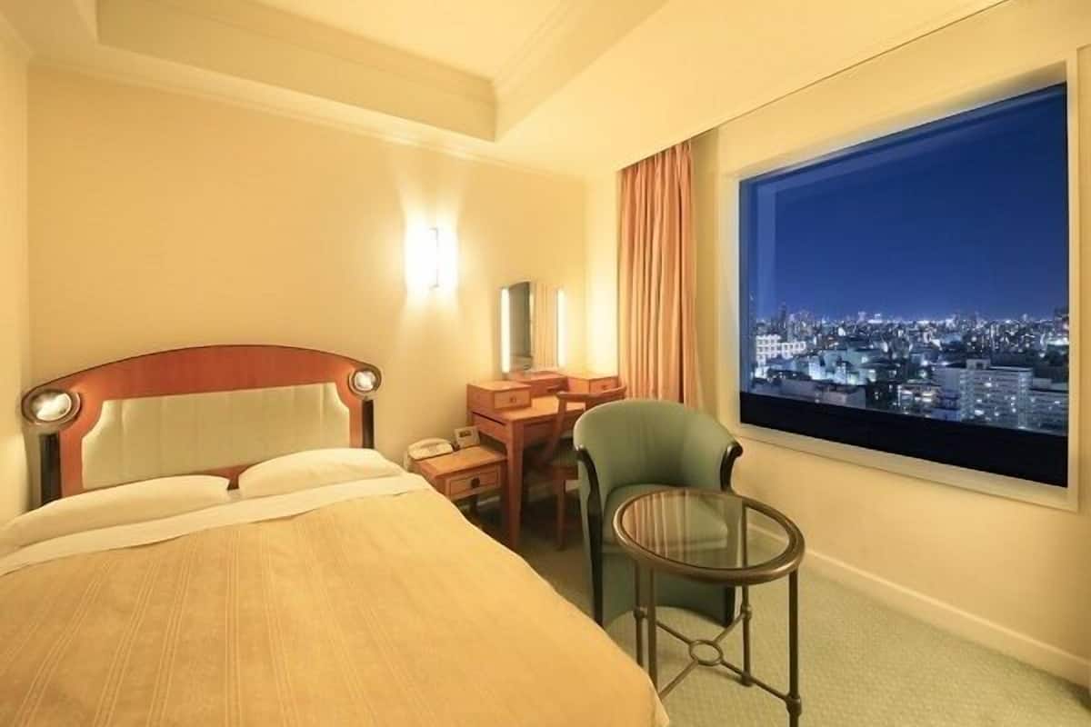Ein Hotelzimmer mit Bett, Schreibtisch, Stuhl, kleinem Tisch und großem Fenster mit Blick auf die Stadt.