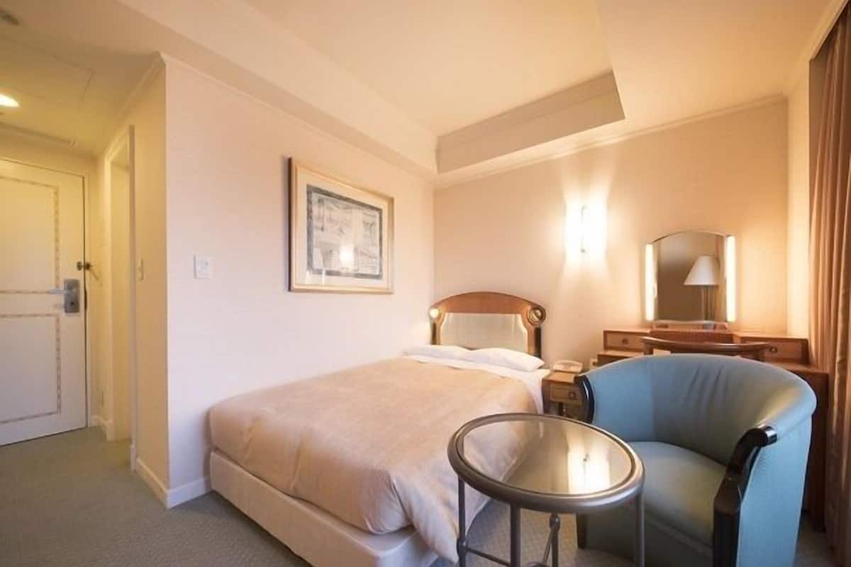Ein Hotelzimmer mit Bett, Sessel, kleinem Tisch, Schreibtisch mit Lampe und einem gerahmten Bild an der Wand.