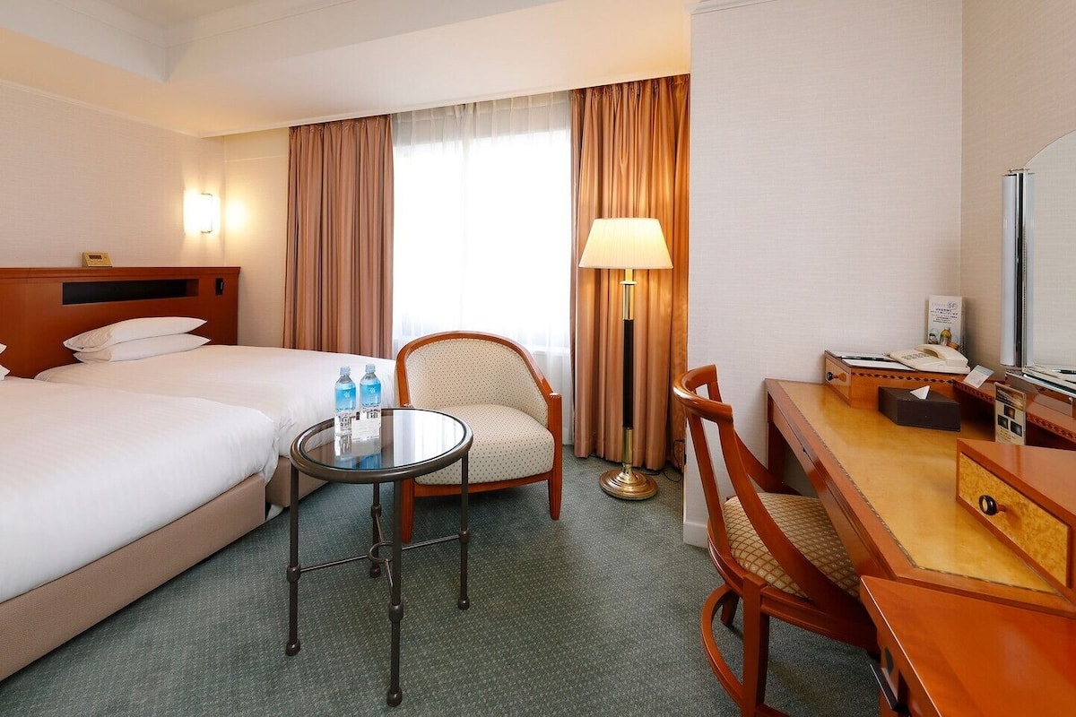 Ein Hotelzimmer mit zwei Betten, einem runden Tisch, einem Stuhl, einem Schreibtisch mit Telefon und einer Lampe.