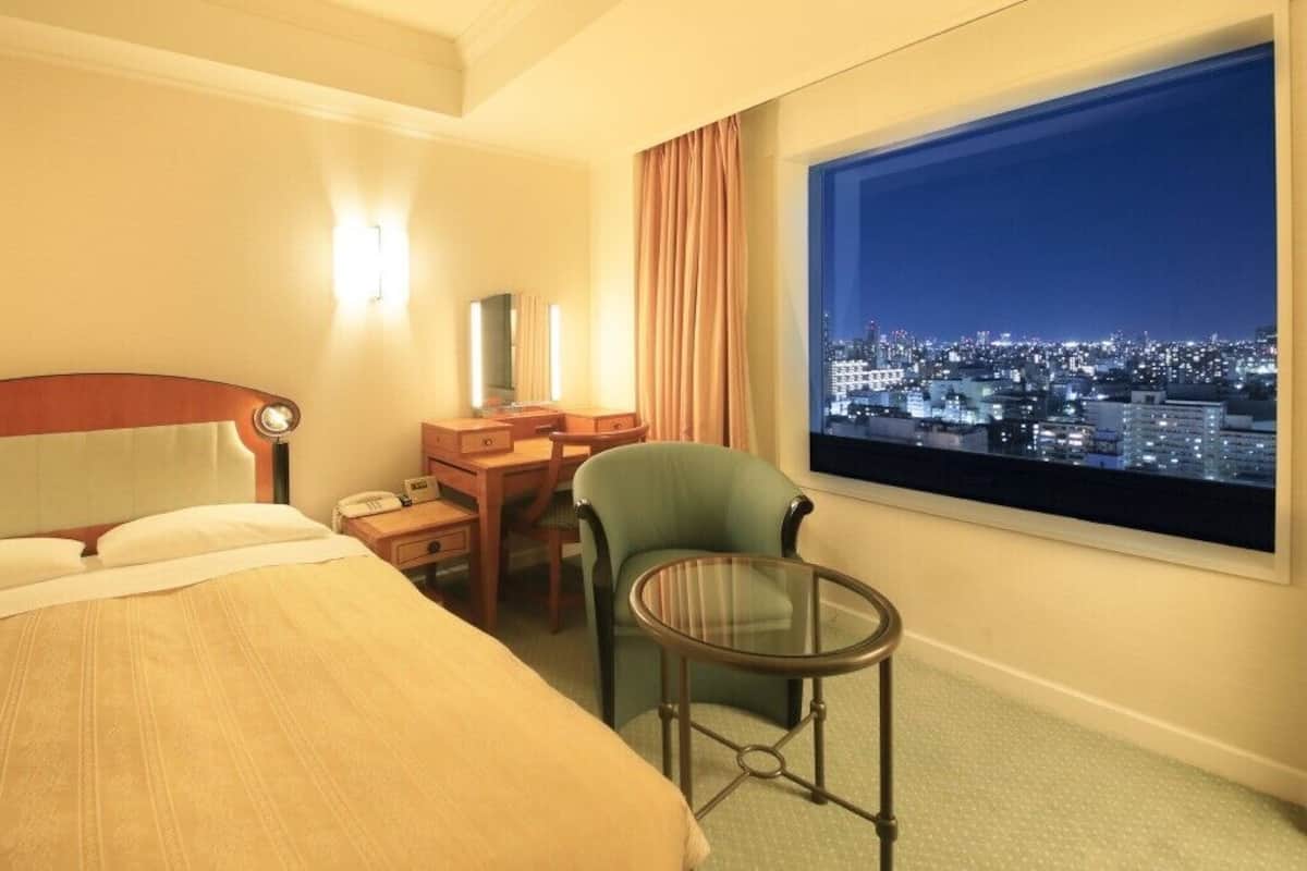 Ein Hotelzimmer mit Bett, Schreibtisch, Stuhl, Fernseher und Fenster mit Blick auf die Stadt.