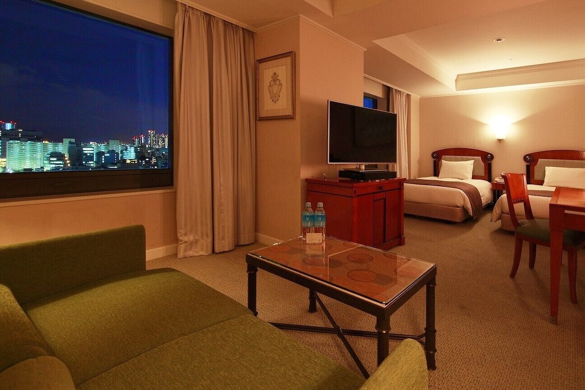 Ein Hotelzimmer mit Blick auf die Stadt, inklusive Fernseher, Schreibtisch mit Lampe, zwei Betten und einem grünen Sofa.